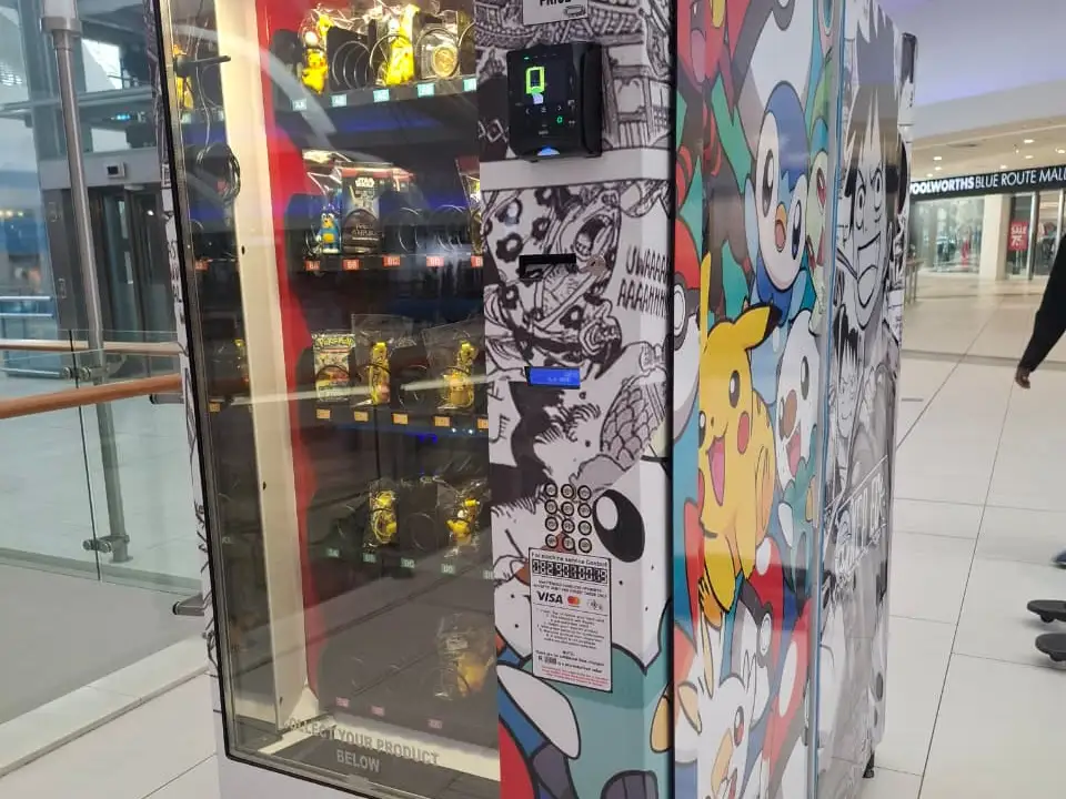 A collector vending machine wrapped in anime artwork, stocked with character figurines, on a shopping mall concourse