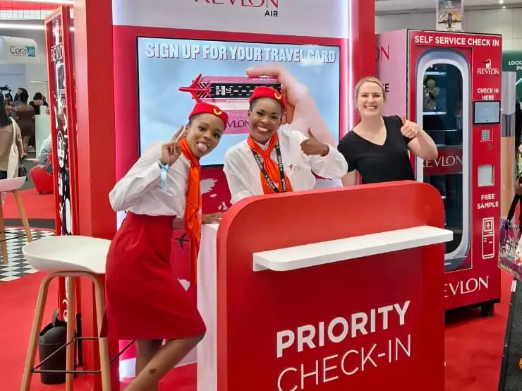 A Revlon Air lead-capture vending machine at a beauty expo, with brand promoters at a priority check-in desk and a free-sample machine beside them