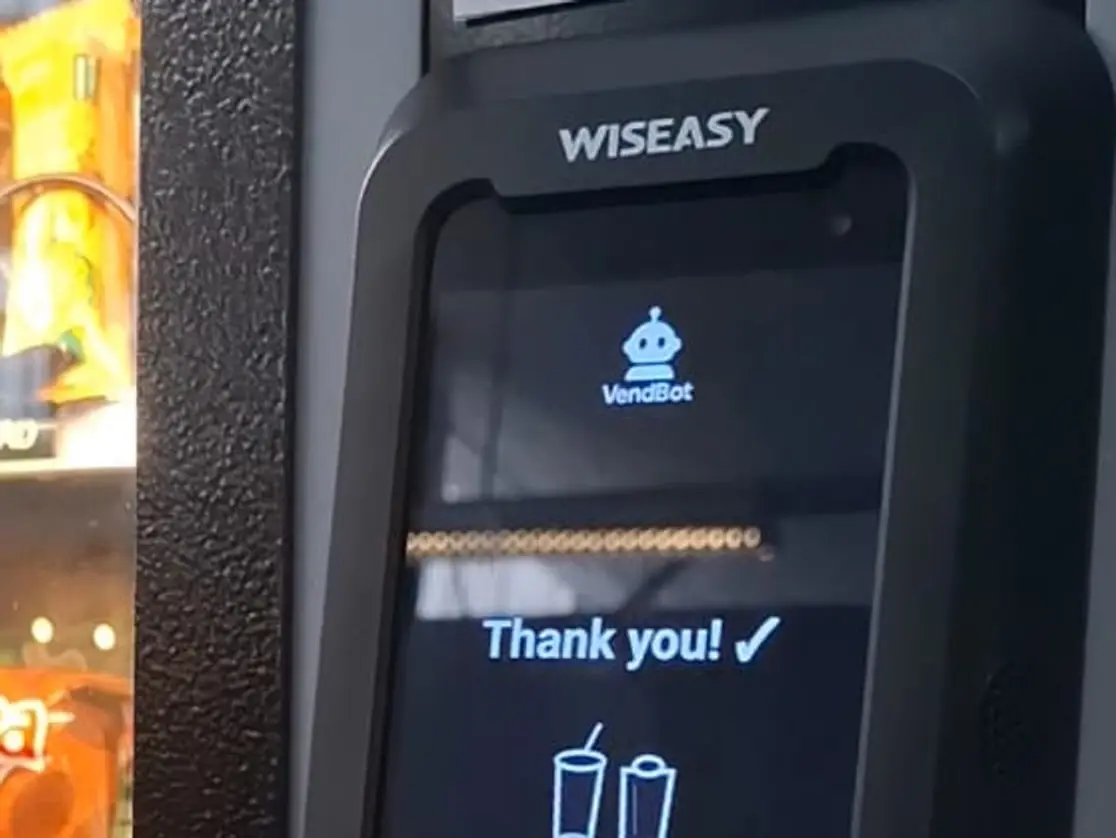 The VendBot P5L card terminal mounted on a vending machine, its screen showing the VendBot logo and a completed sale, with stocked shelves behind