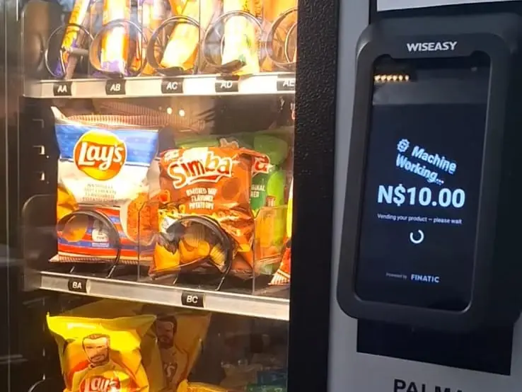 The same terminal mid-vend, its screen reading N$10.00 in Namibian dollars above 'Vending your product — please wait' and 'Powered by Finatic', with shelves of crisps behind the glass