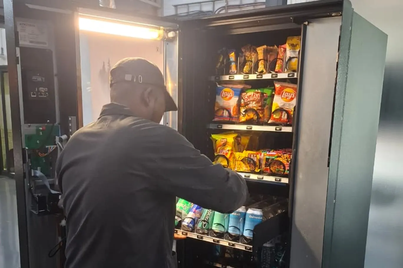An operator restocking a vending machine, its door open and the shelves filled with crisps and cold drinks